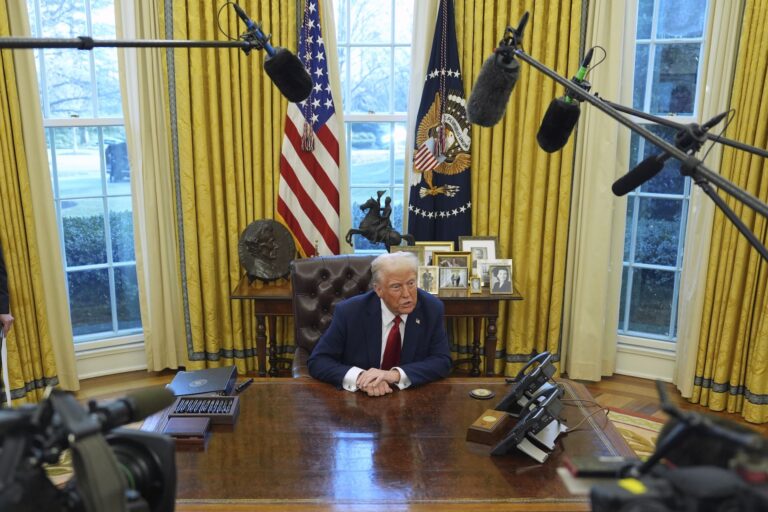 A high-angle shot of Donald Trump sitting at the Resolute Desk in the Oval Office, with the gold-hued curtains open behind him. He's speaking to reporters, and there are boom mics hovering in the foreground.