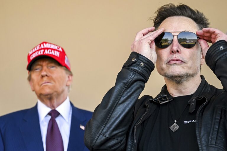 Elon Musk putting on sunglasses, with Donald Trump looking on in the background. Musk is wearing a black leather jacket. Trump his wearing a blue suit, a red "MAGA" hat.