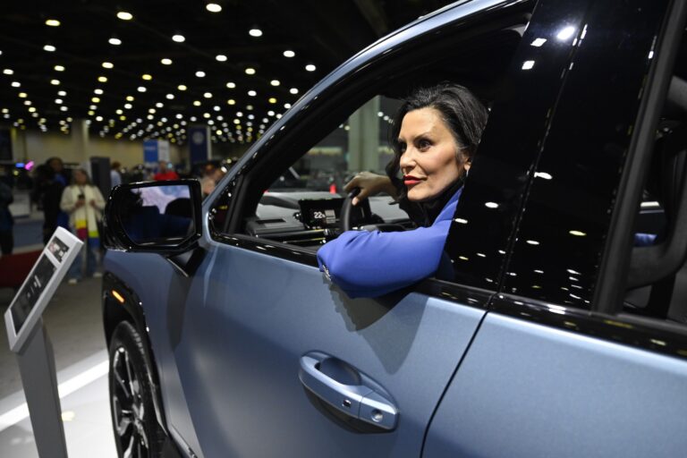 A sideview shot of a blue-grey pickup with Michigan Governor Gretchen Whitmer in the driver's seat. The truck is on display in a convention centre. Its driver's-side window is open and Whitmer, who is wearing a bright blue suit jacket, is resting her elbow on the door frame.