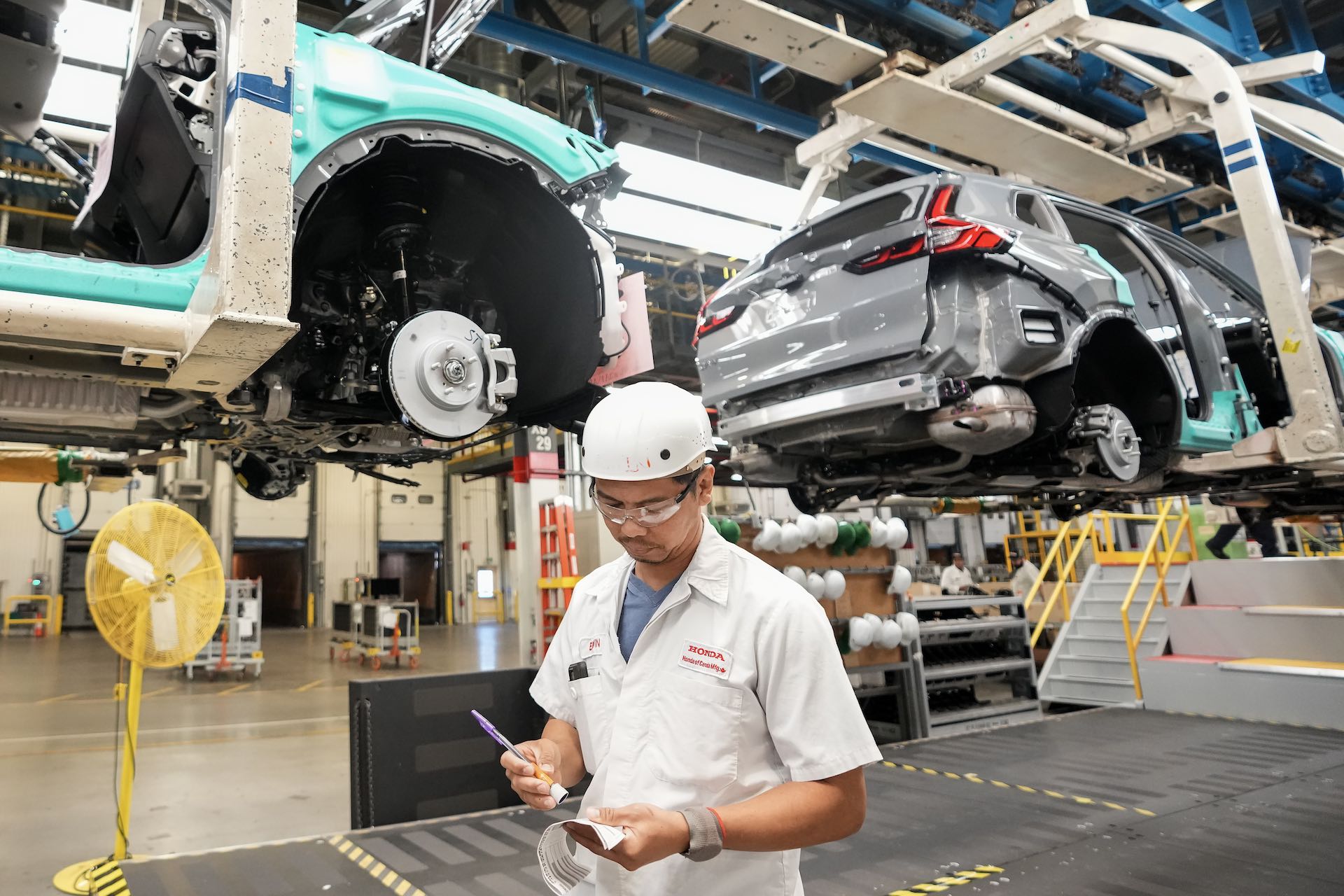 A worker in a white short-sleeve work shirt and white hardhat stands below factory gantries carrying partially assembled bodies of small SUVs. He's wearing safety glasses and marking a piece of paper with a pen.