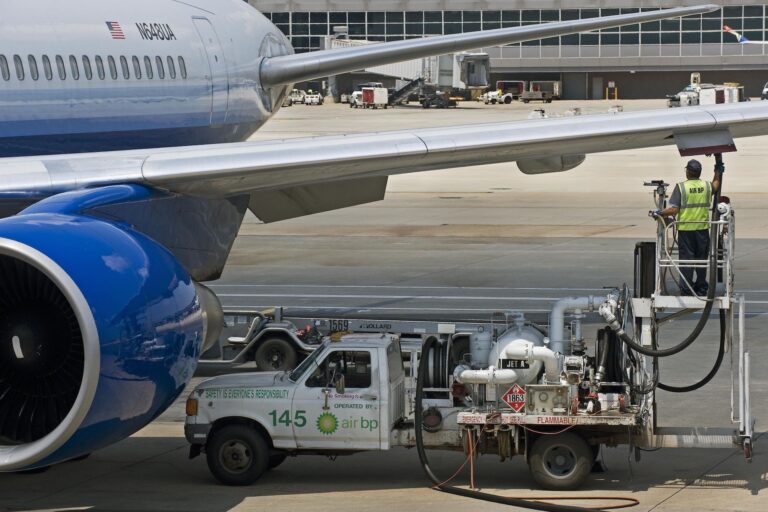 A view of a worker in a reflective vest under the wing of a United Airlines jet, connecting a fuel line from the platform of a white pumping truck.