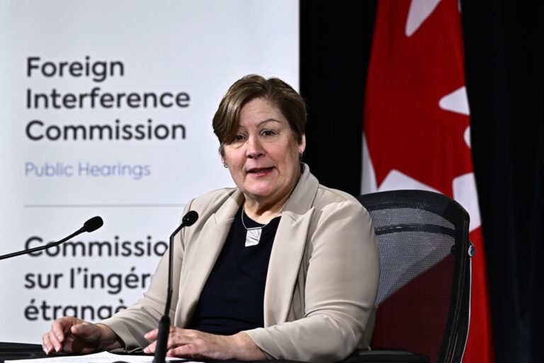Marie-Josee Hogue sits on a desk chair and speaks into two small desktop microphones. She has short, brown hair and a beige blazer, and sits at desk in front a of Canadian flag and a projected backdrop that says "Foreign Interference Commission Public Hearings."