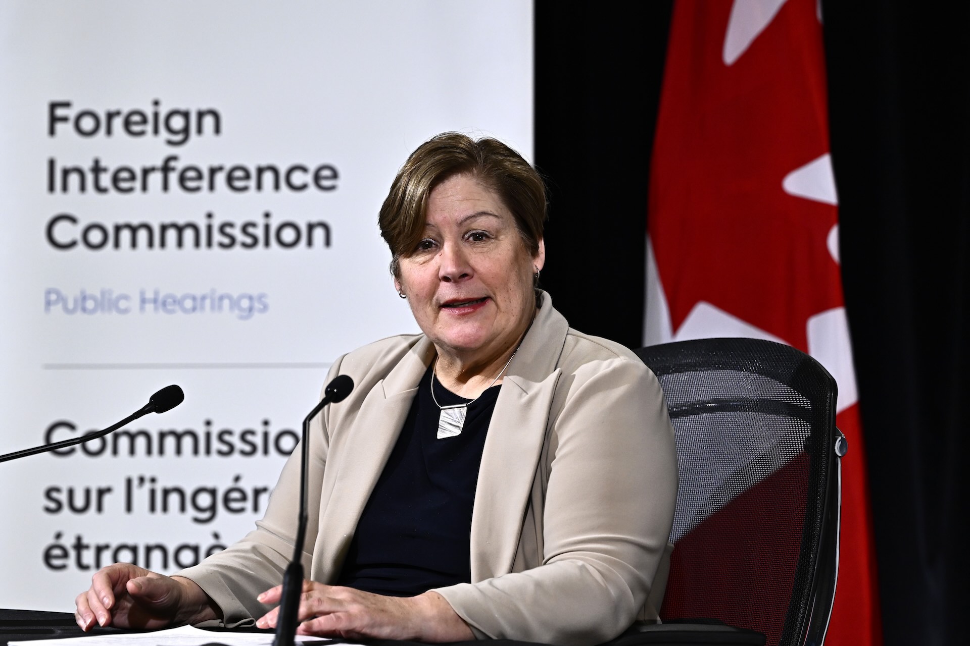 Marie-Josee Hogue sits on a desk chair and speaks into two small desktop microphones. She has short, brown hair and a beige blazer, and sits at desk in front a of Canadian flag and a projected backdrop that says "Foreign Interference Commission Public Hearings."