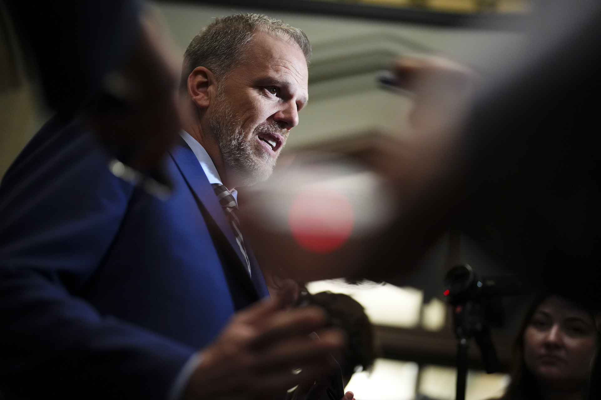 A low-angle profile shot of Mark Holland in a media scrum, behind a foreground of blurred hands and cameras. He is wearing a blue suit jacket and tie, and has a concerned expression on his face.
