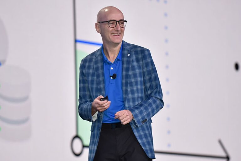Neil Cawse stands in front of white screen, holding a small remote in his hand