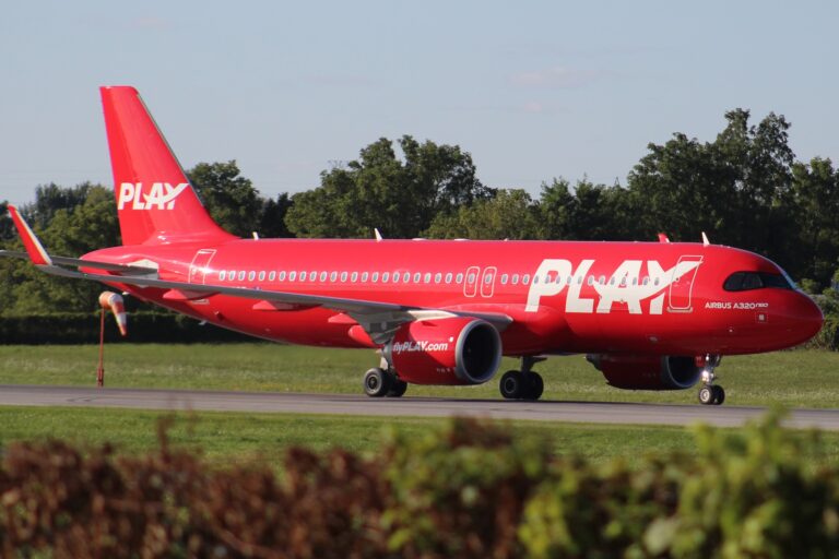A red Play Airlines Airbus A320neo on ground with green bushes and trees around.