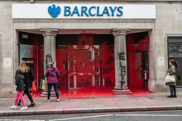 An image of a Barclay's bank entrance covered in red paint with "Elbit" spray-painted in black on its decorative columns. Several pedestrians have stopped to look.