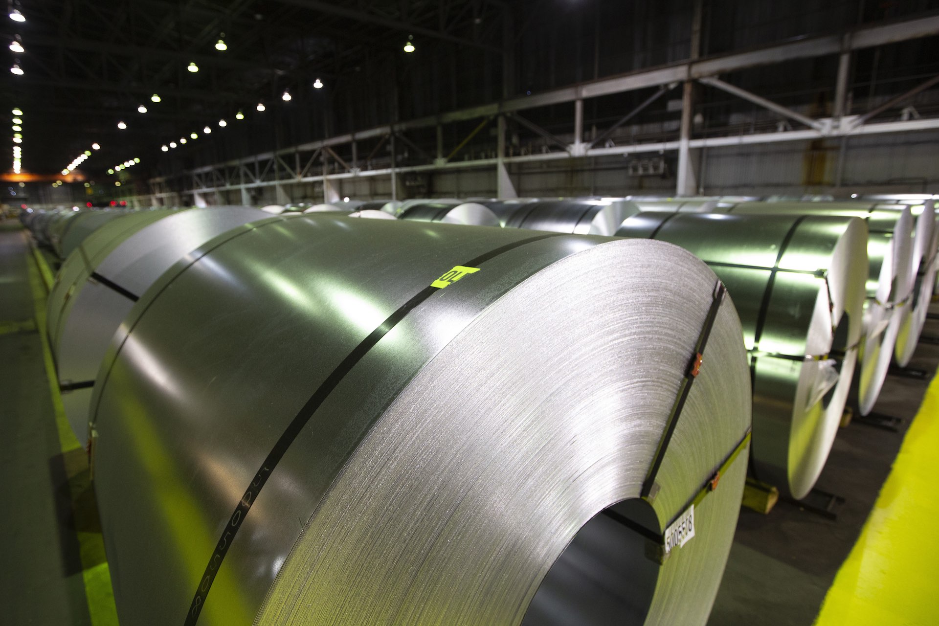 A shot of large coils of thin, shiny steel waiting in rows for shipping, in a large warehouse. The rows reach from the foreground to deepest background of the frame.