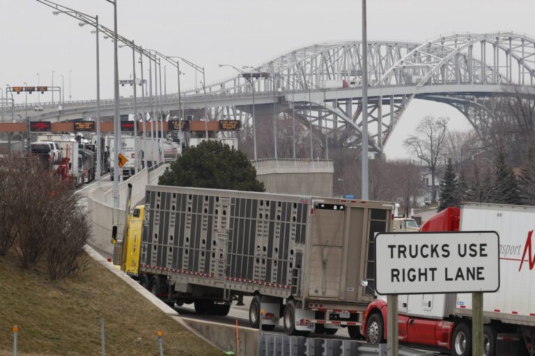 A snarl of transport trucks on the ramp toward a large metal bridge, whose iron arches appear in the background. A border checkpoint with illuminated signage over each lane is visible on the upper left side of the frame.