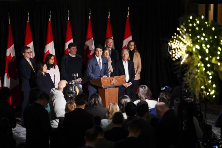 Justin Trudeau speaks from a wooden lectern embossed with a carved maple leaf. He is wearing a blue suit and behind him are members of his cabinet. There is a row of seven Canadian flags in the background.
