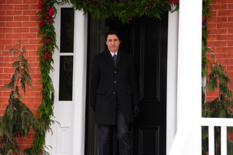 Justin Trudeau appears at the entrance to Rideau Cottage in a dark three-quarter length jacket and blue tie. The black door to the red brick home is wreathed with festive boughs and holly berries.