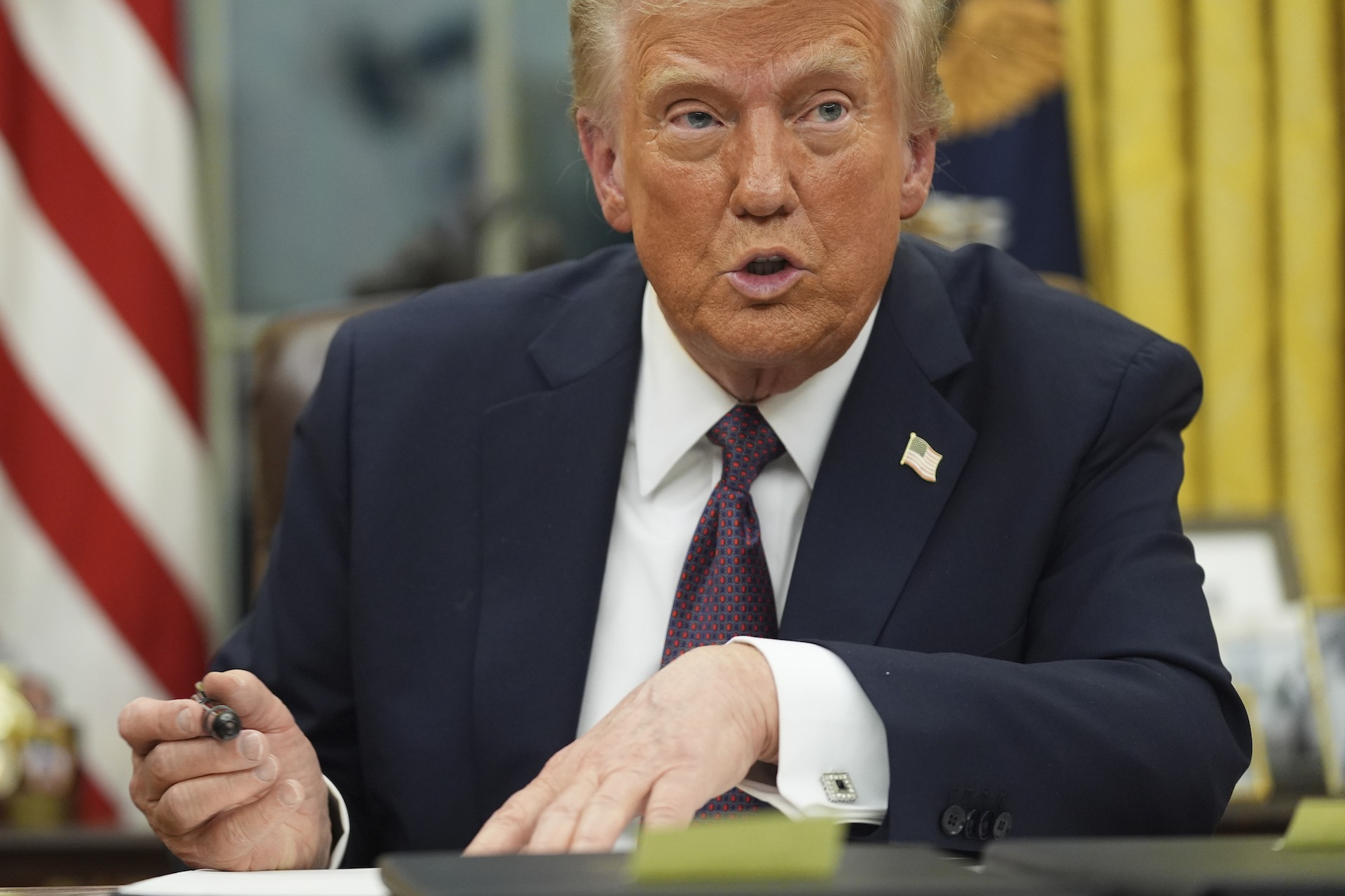 A close-up of Donald Trump at the Oval Office desk with a large pen poised over a document. Trump is wearing a blue suit jacket with an American flag pin on the lapel. He is in mid-sentence. The orange hue of his face contrasts sharply with the white skin of his hands.