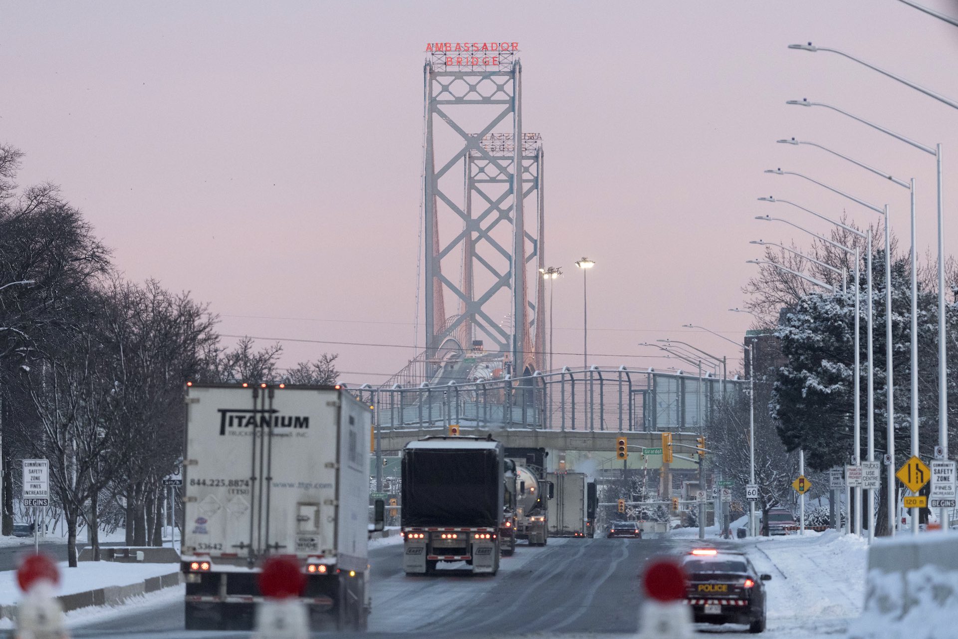 A view of the iconic bridge leading from Windsor, Ont. to Detroit, shot from the Canadian side. The angle shows transport trucks headed toward the bridge, whose towering steelwork and and red neon sign reading "Ambassador Bridge" are visible in the distance.