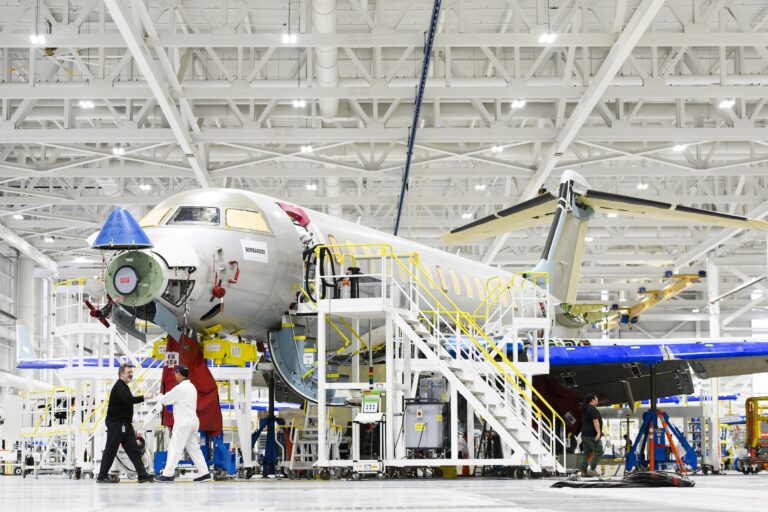 A partly assembled airplane sits in a hangar, surrounded by stairs and scaffolding. Four workers can be seen around the plane and in the background.