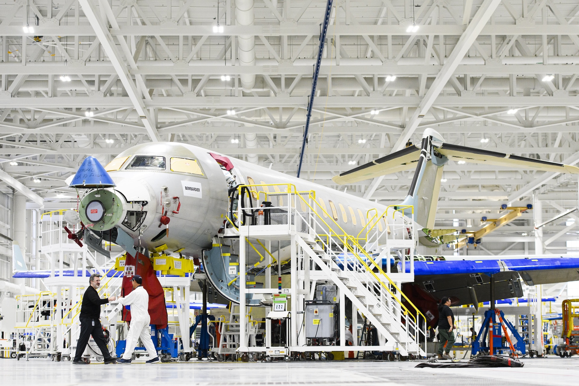 A partly assembled airplane sits in a hangar, surrounded by stairs and scaffolding. Four workers can be seen around the plane and in the background.