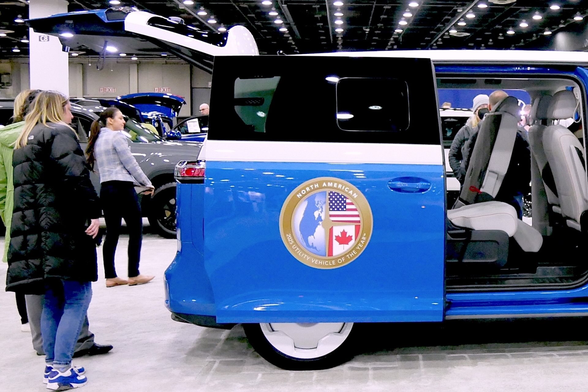 A blue van on a car show floor with its side and rear doors open. The side door has a circular logo with the U.S. and Canadian flags on it proclaiming it the North American 2025 Utility Vehicle of the Year.