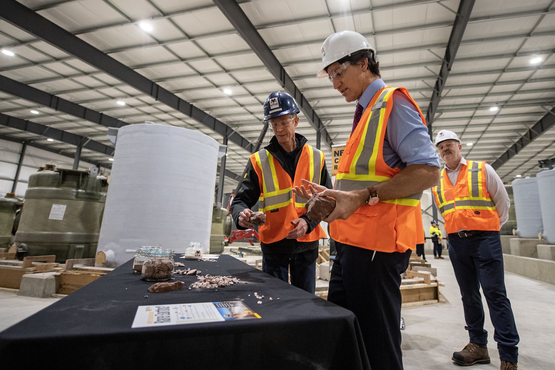 Three men including Prime Minister Justin Trudeau wear construction vests, hard hats and safety glasses indoors in a processing plant. Justin Trudeau holds a rock and appears to be conversing with the man next to him, also holding a rock, about the various minerals on the table in front of them.