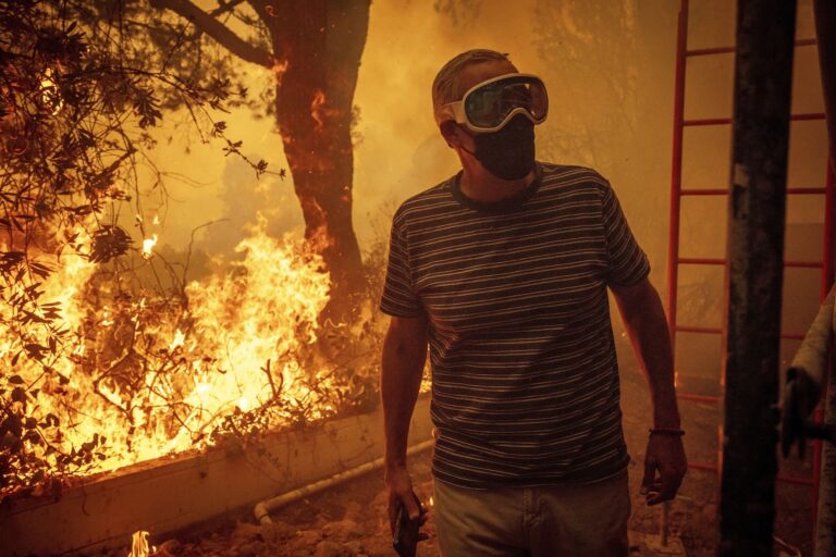 A man wearing a mask and goggles stands near the side of a building as large flames consume trees and bushes behind him in the Pacific Palisades neighbourhood of Los Angeles in January 2025.