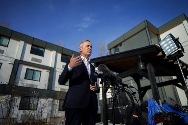 Mark Carney in a dark suit and white open-necked shirt, addressing the media from behind a lectern with mics on it. There is a new-looking lowrise housing complex behind him.