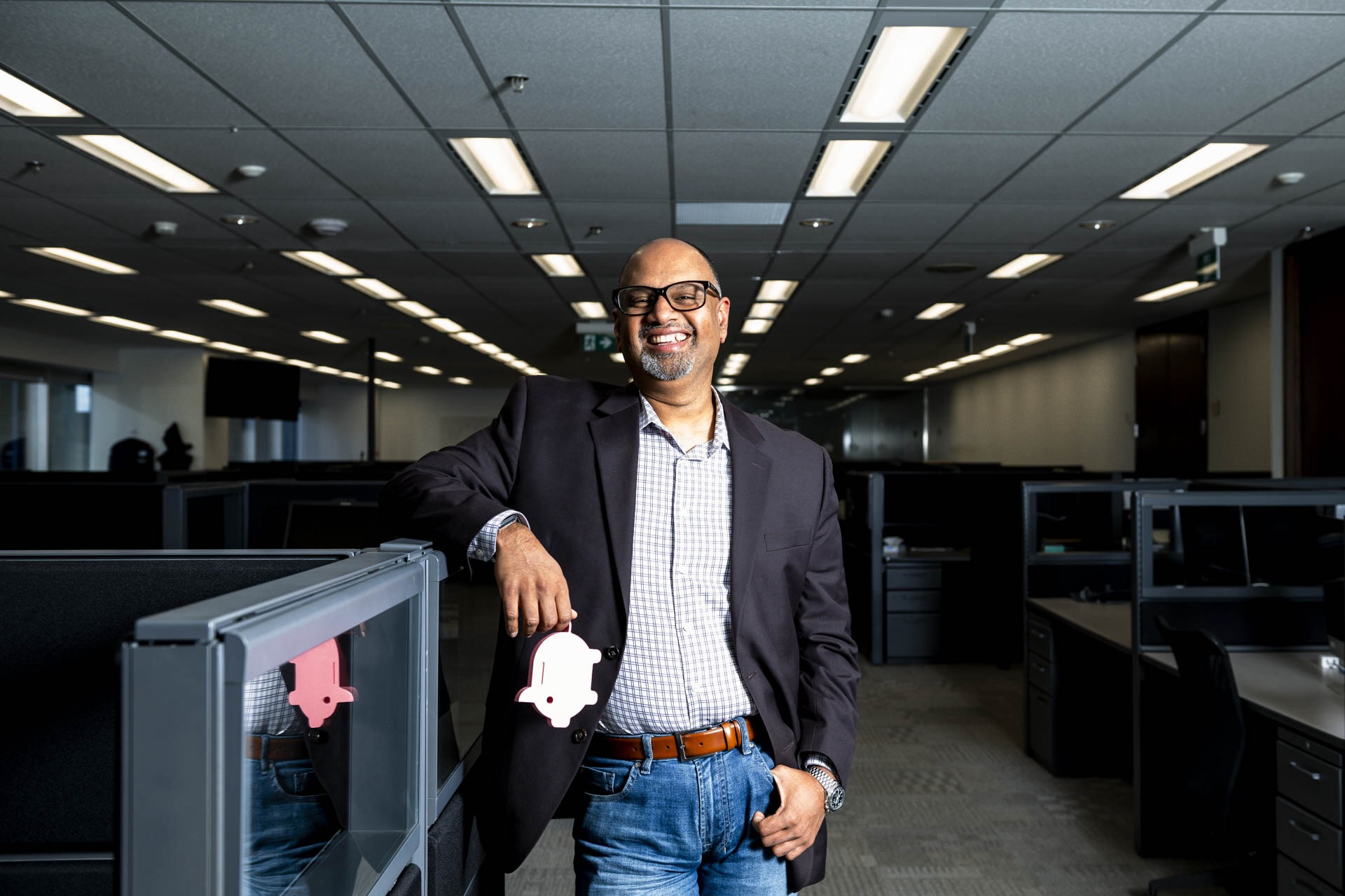 A man in a blazer, jeans and a collared shirt stands in an office, leaning an arm on a cubicle divider and smiling at the camera. He is holding a logo shaped like a piggy bank by the tail.