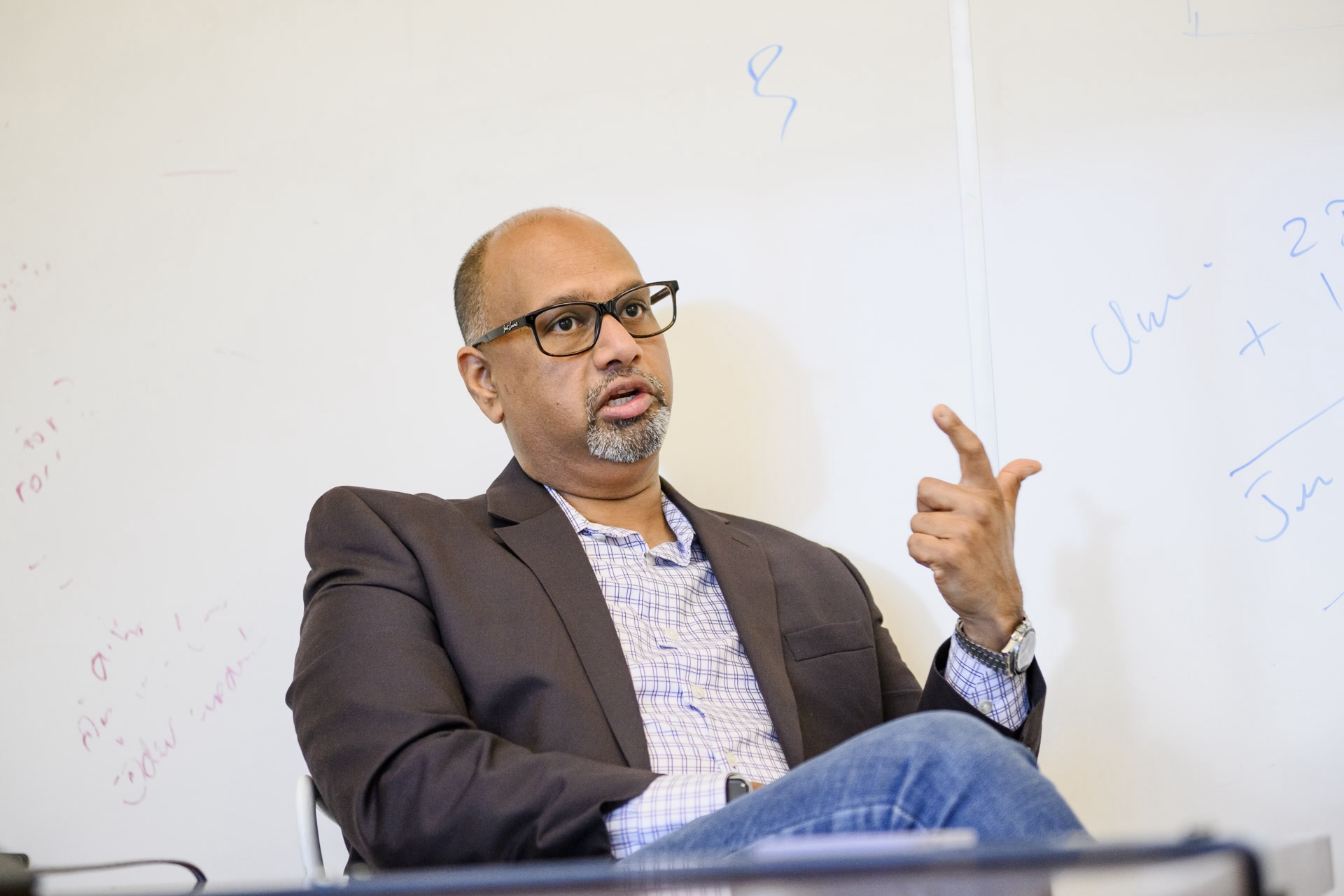 A man in a collared shirt, blazer, jeans and glasses sits in front of a whiteboard with partially erased notes on it. He is speaking and raising his left hand to make a gesture