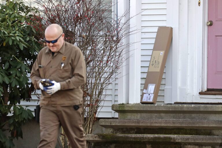 A UPS delivery driver leaving the front step of a white-sided home with a pink door. There is a large rectangular cardboard package leaning against the doorjamb.