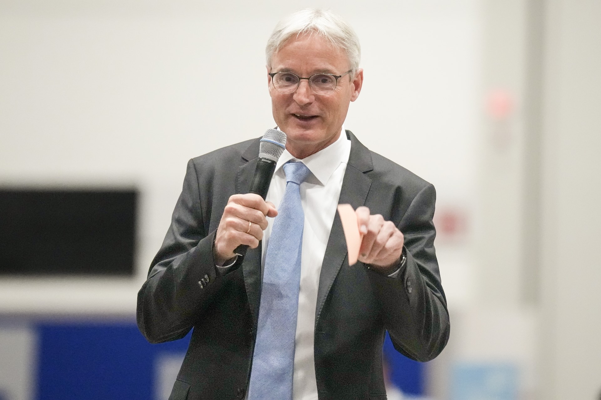 Waist-up picture of a man in a gray suit with white hair and glasses holding a microphone in one hand and gesturing to the camera with a note card as he speaks