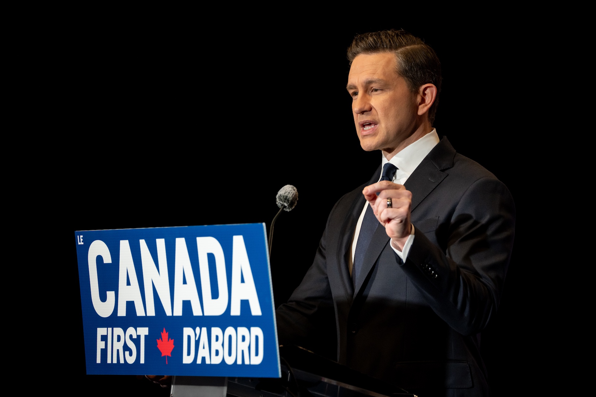 A shot of Pierre Poilievre speaking into a microphone at a lectern, against a black background. He's wearing a dark suit and there's a sign on the lectern reading "Canada First."
