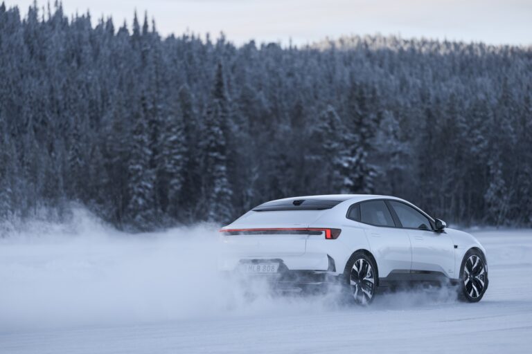 A sleek white vehicle driving across a frozen lake, with a backdrop of trees and snow billowing behind it.