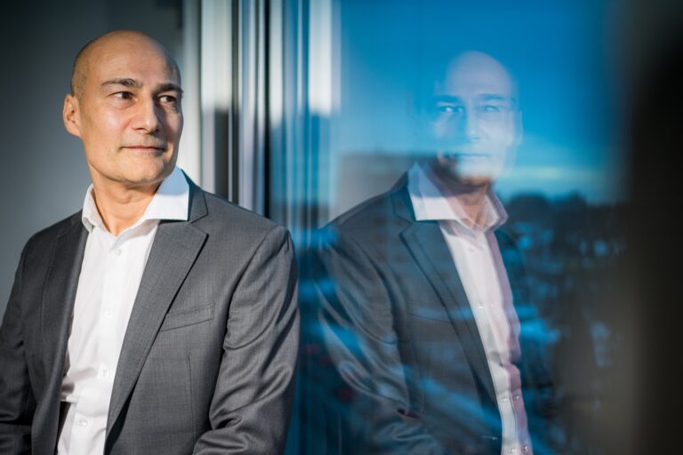 A head-and-shoulder shot of Rajiv Gupta seated next to a window that gives a blurred reflection of his image. Gupta's head is shaven. He's wearing a grey blazer and white open-necked shirt.