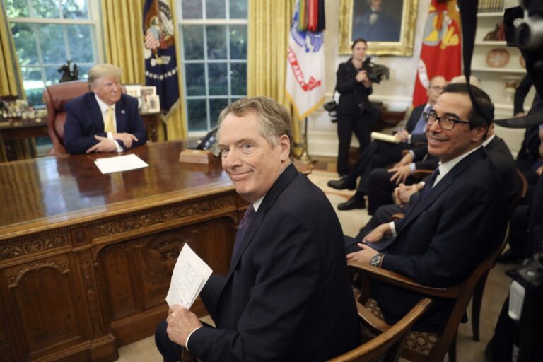 A picture in the Oval Office with a slightly out-of-focus Donald Trump at the desk and a row of men seated in chairs in front of him and photographers standing nearby. The closest man in a chair has turned to smile at the camera