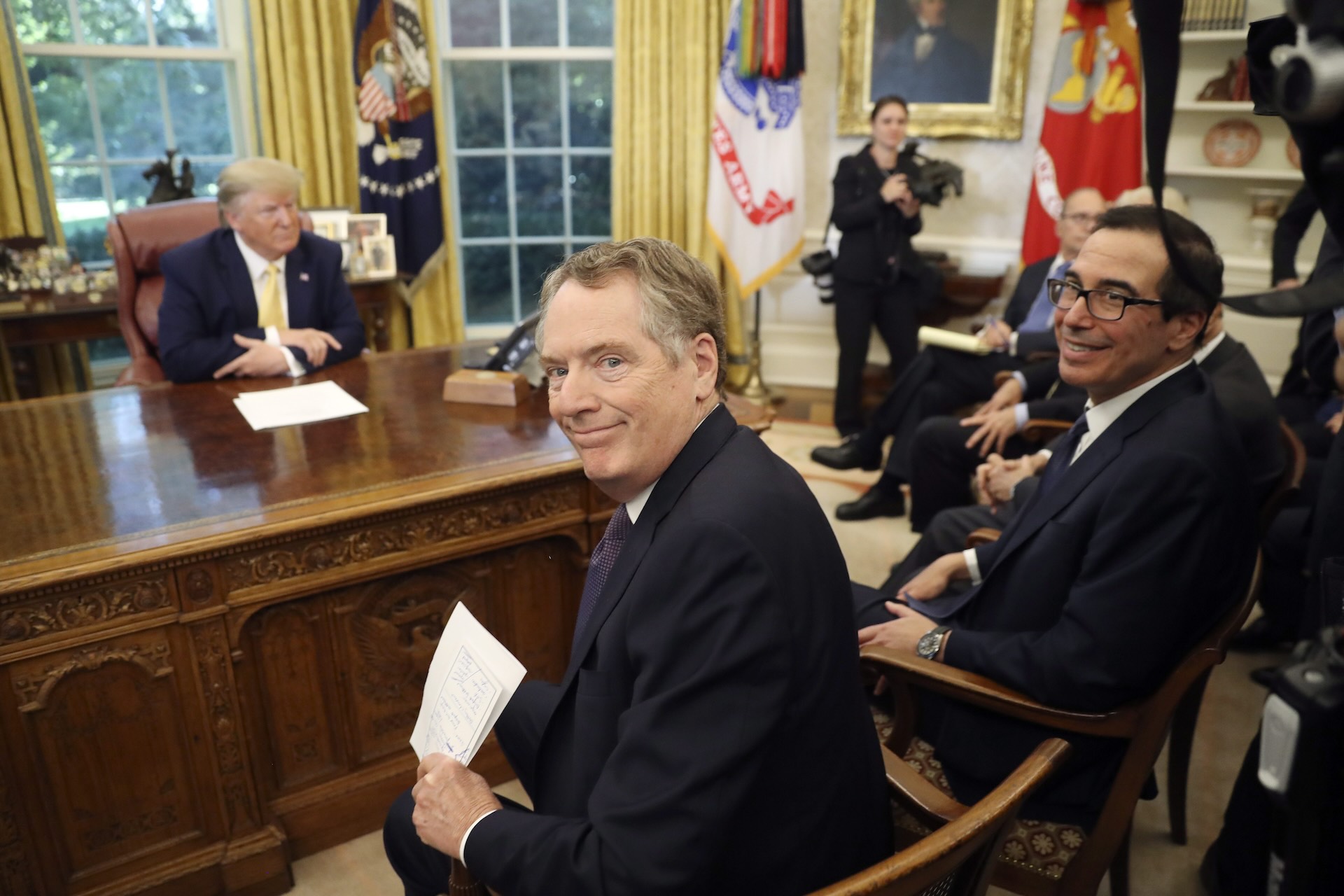 A picture in the Oval Office with a slightly out-of-focus Donald Trump at the desk and a row of men seated in chairs in front of him and photographers standing nearby. The closest man in a chair has turned to smile at the camera