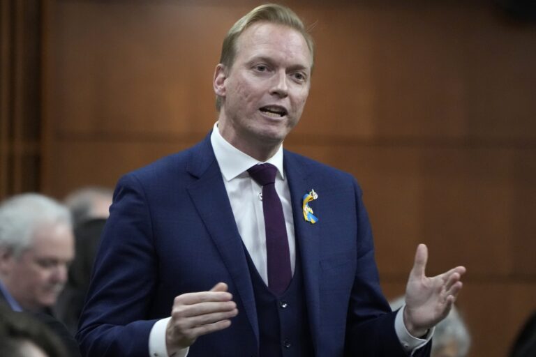 A head-and-shoulder shot of MP Ryan Williams standing and speaking in the House of Commons. He's wearing a blue jacket with a blue-and-yellow lapel ribbon, and a purple tie