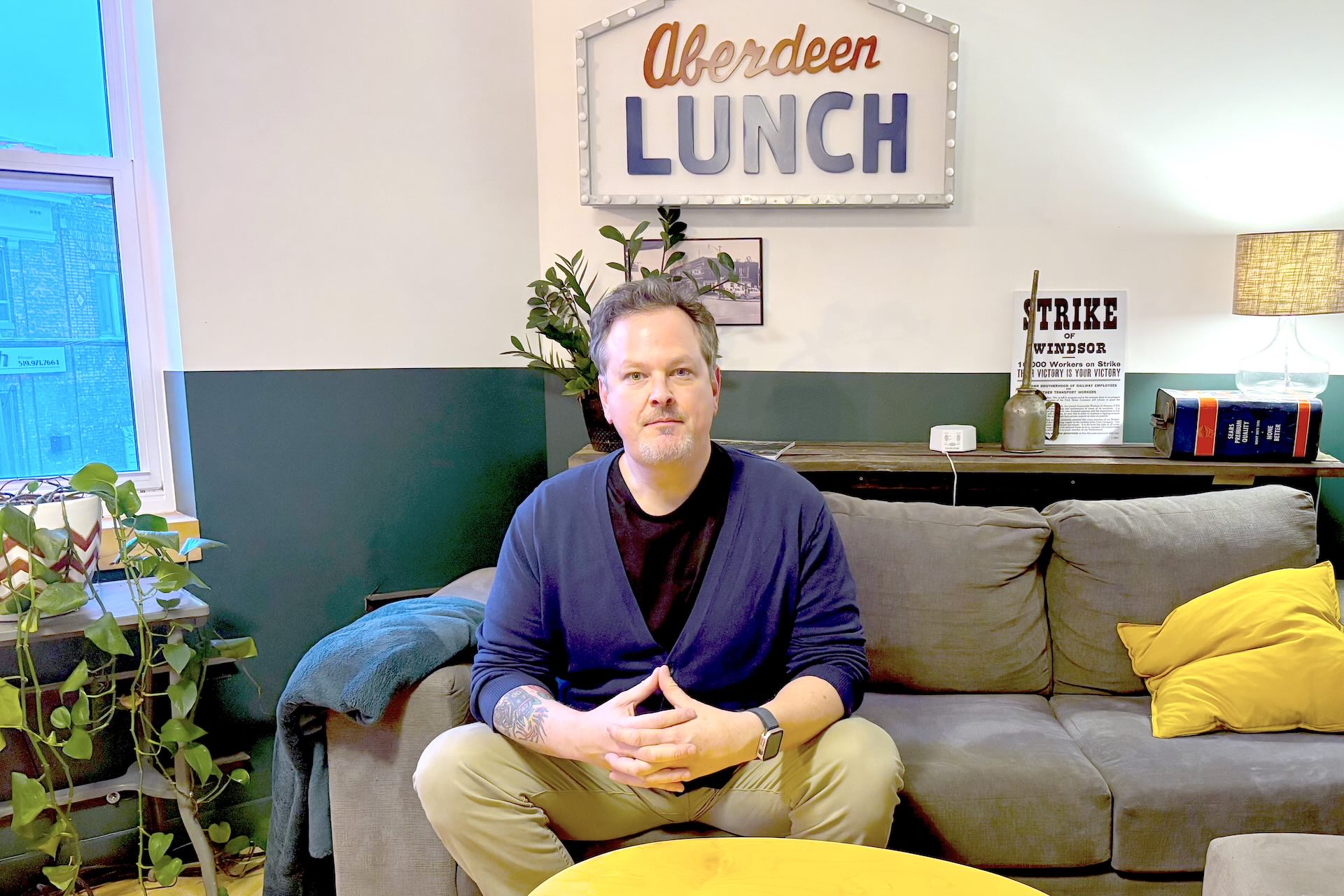 A man in a blue sweater sits on a couch looking at the camera. A sign on the wall above him says "Aberdeen Lunch."