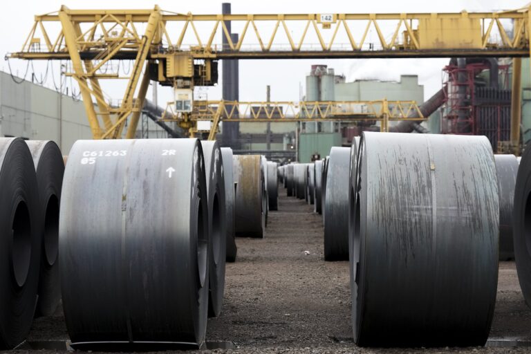 Dark rolls of steel sit in rows, against a backdrop of yellow gantries and steel mill buildings.