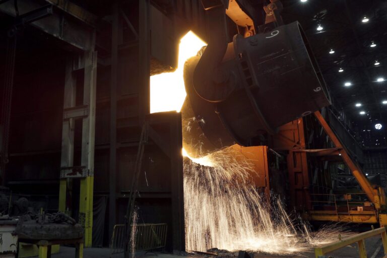 Molten metal pours from a massive cauldron into a furnace, as a cascade of sparks flows from the opening of the furnace onto the floor of a steel mill.