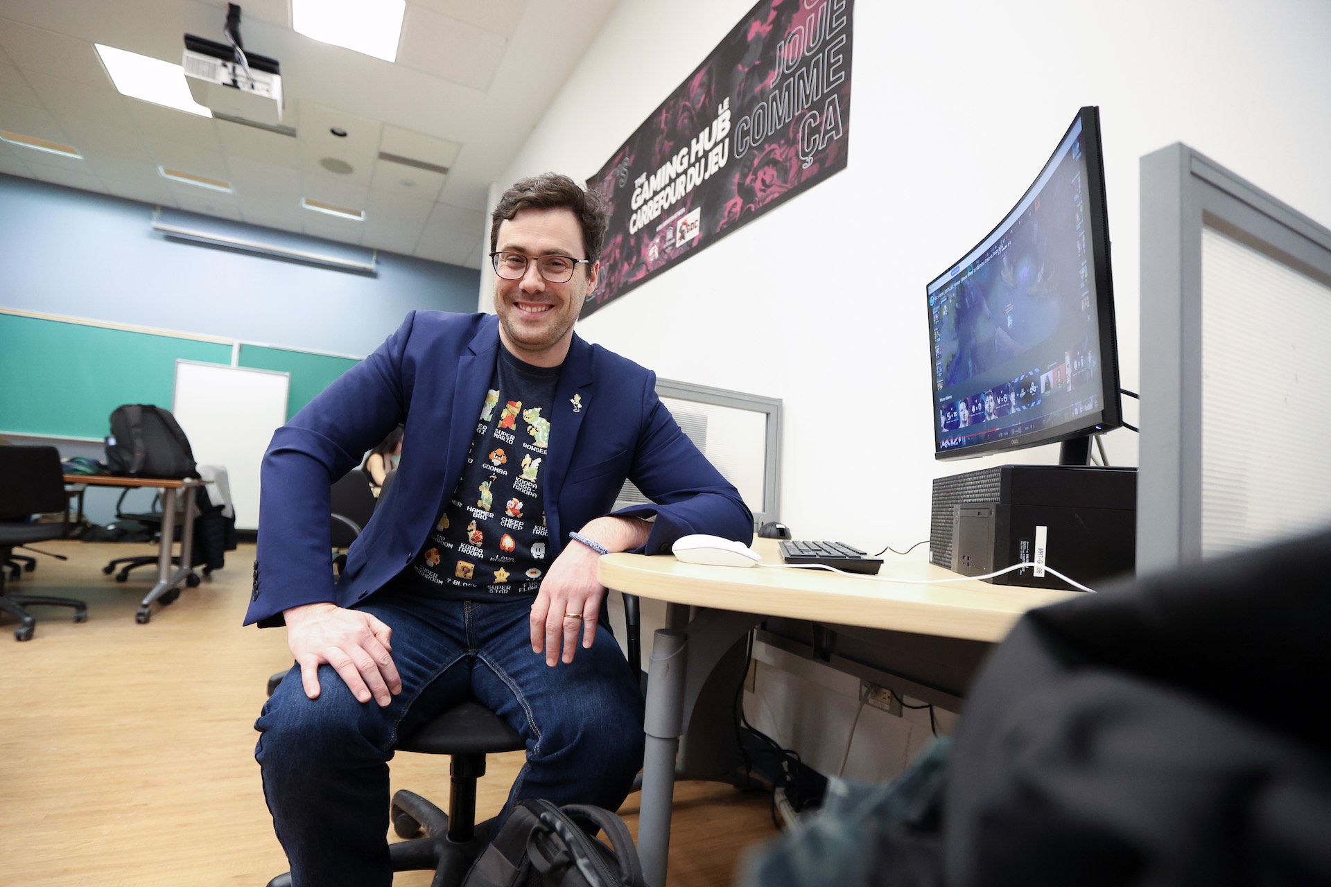 Thomas Burelli, wearing a blue blazer over a t-shirt with video gaming icons on it, sits smiling at the camera beside a curved video monitor. There is a black-and-purple banner on the wall that reads "The Gaming Hub."