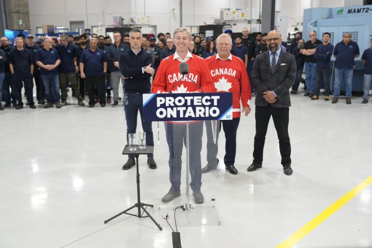 Tim Houston stands at a lectern wearing a red Team Canada hockey jersey. Behind him, just to his left, is Ontario PC Leader Doug Ford. There is a crowd of factory workers looking on from the background, and a sign on the lectern reading "Protect Ontario."