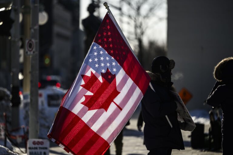A protestor in Ottawa holds a flag with a Canadian flag on one side and an American flag on the other. The protester is in silhouette, but light shining through the flag makes the US side of the flag visible through the Canadian side.