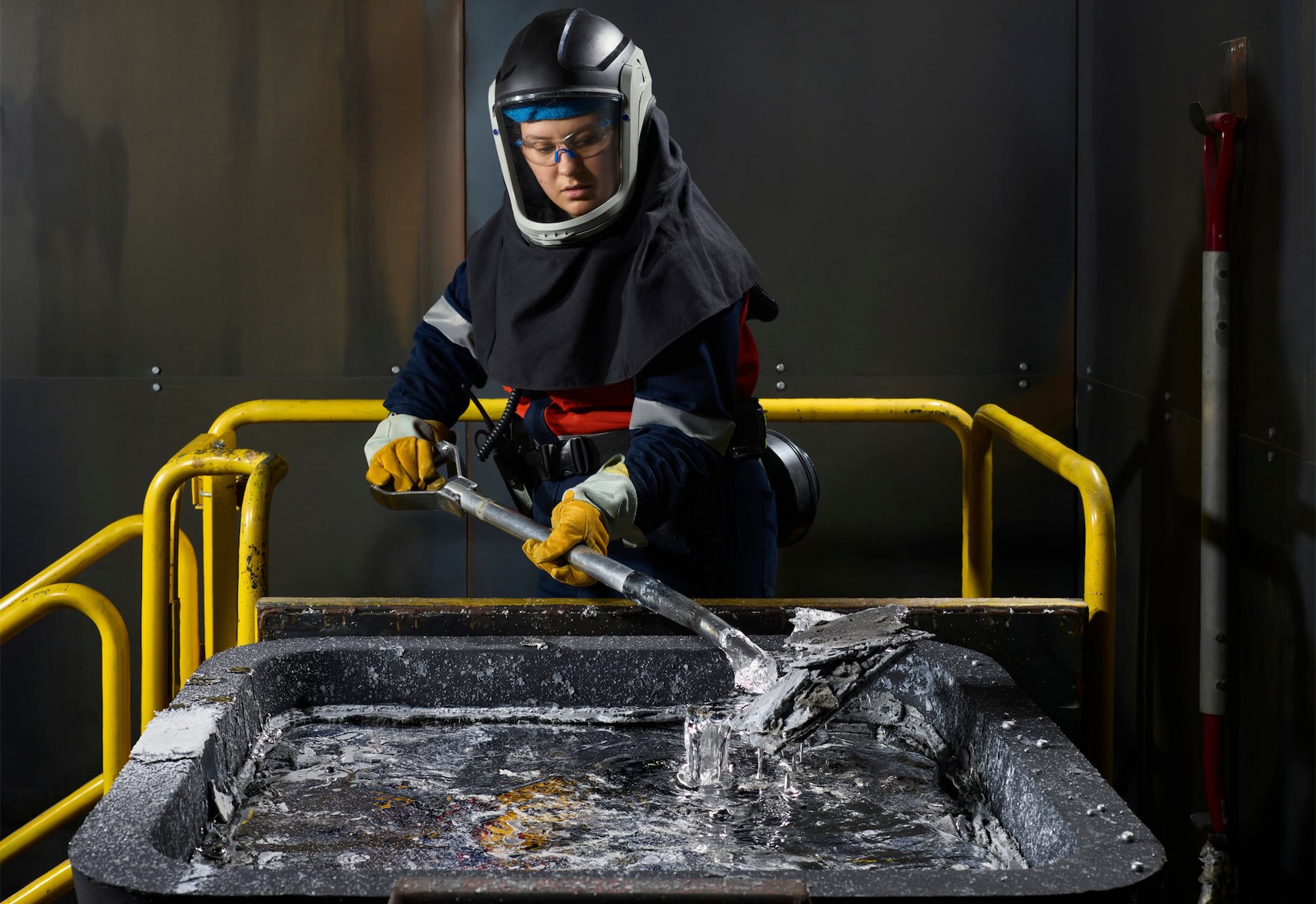 A person wearing protective gear uses a tool to manipulate liquid metal in a large, rectangular container surrounded by yellow railings in a workshop.
