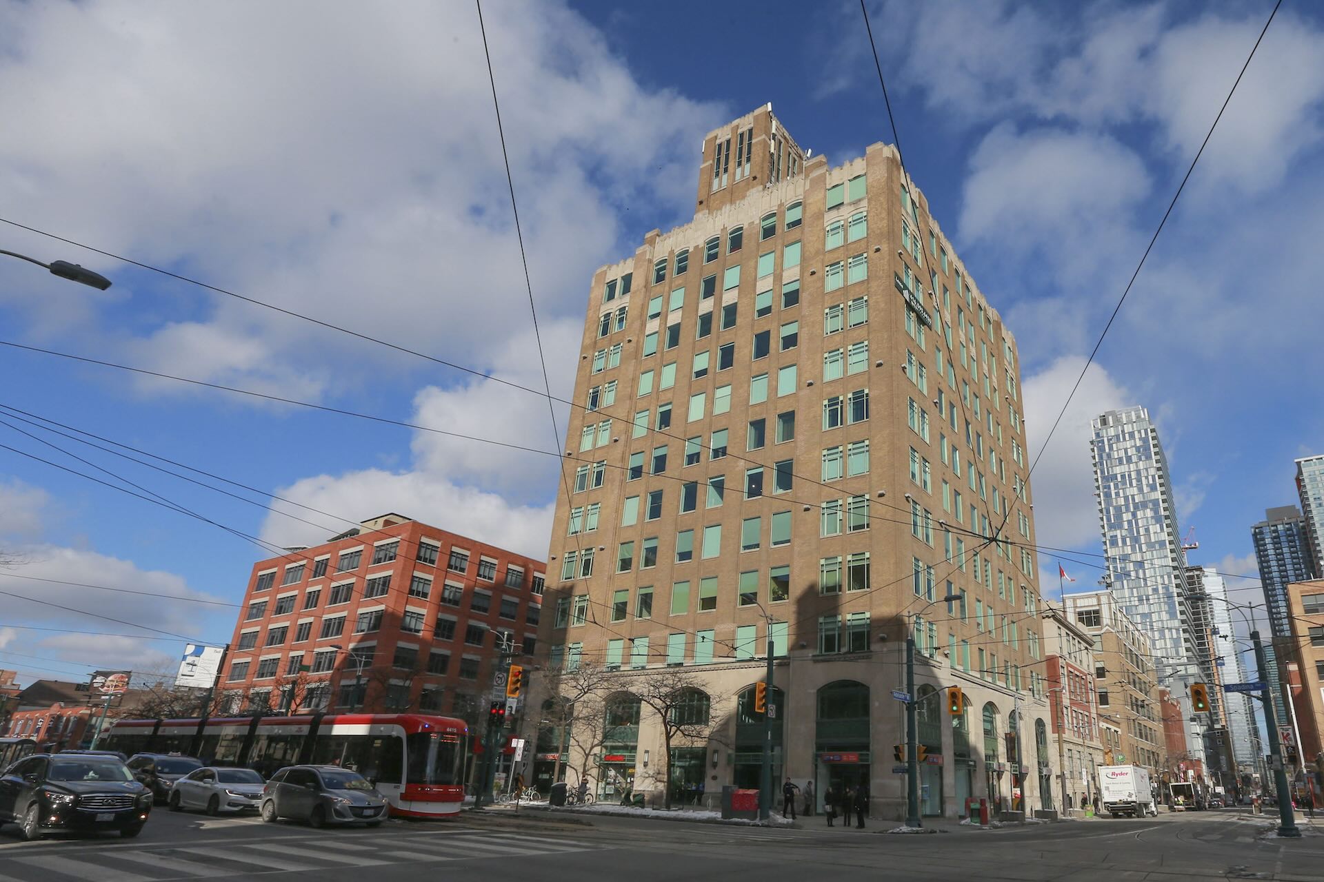 View of brown art-deco building on a Toronto city street scene, surrounded by buildings as a streetcar, and cars pass by at intersection.