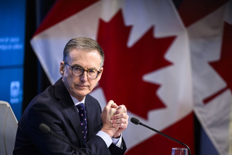 Bank of Canada governor Tiff Macklem in a suit and glasses sitting at a table with Canadian flags in the background.