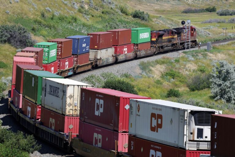 Freight train with colorful shipping containers travels through a grassy, hilly landscape on a clear day.