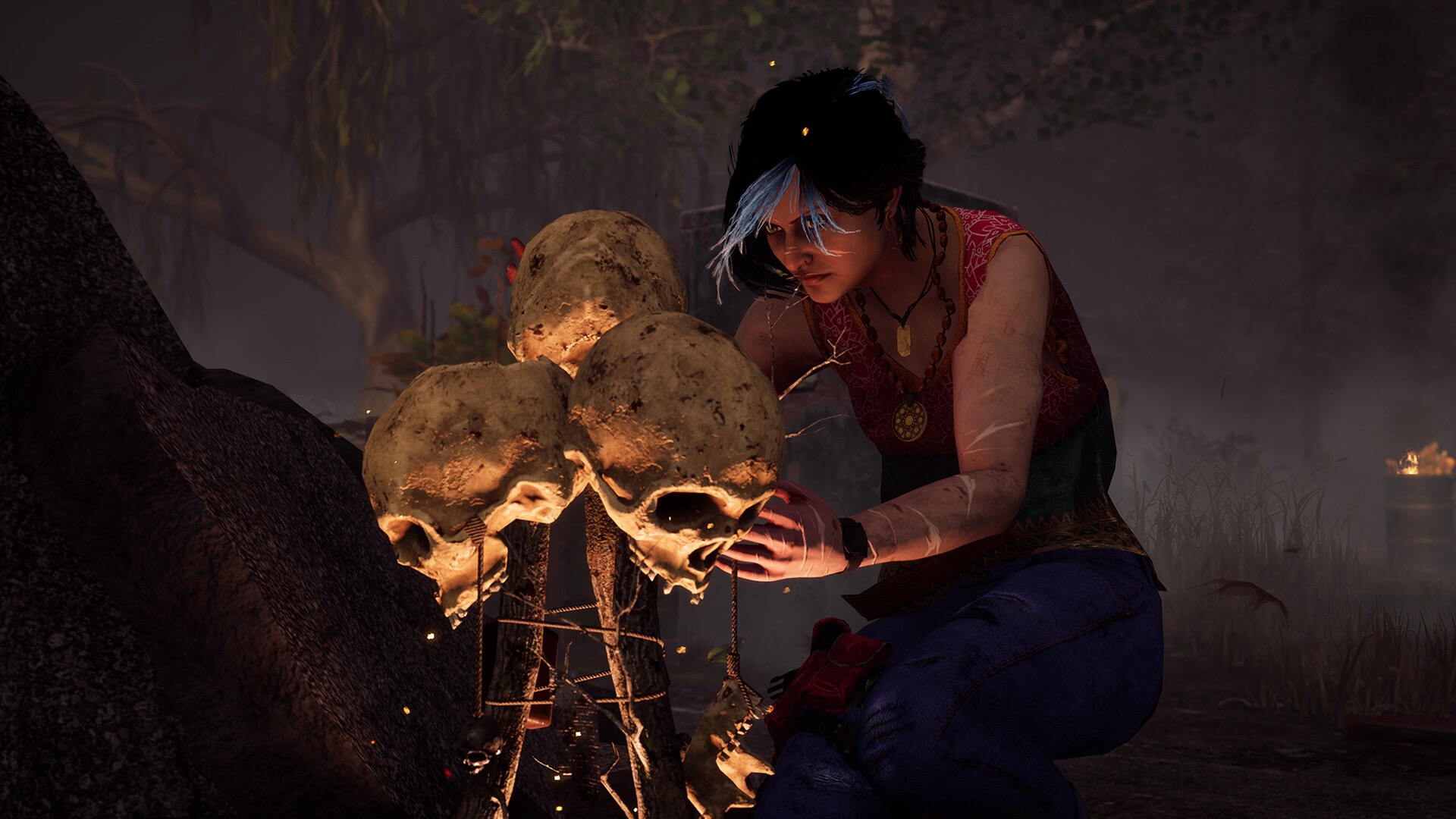 A woman in a red vest and an amulet around her neck crouches down in the woods to examine a small construction made of three skulls and various branches tied together.
