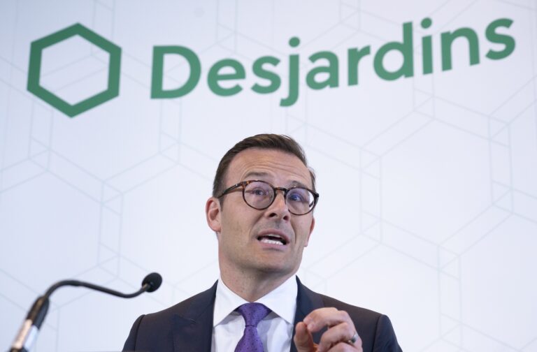 Desjardins president and CEO Guy Cormier gives a speech on stage at a news conference featuring a company banner in the background.