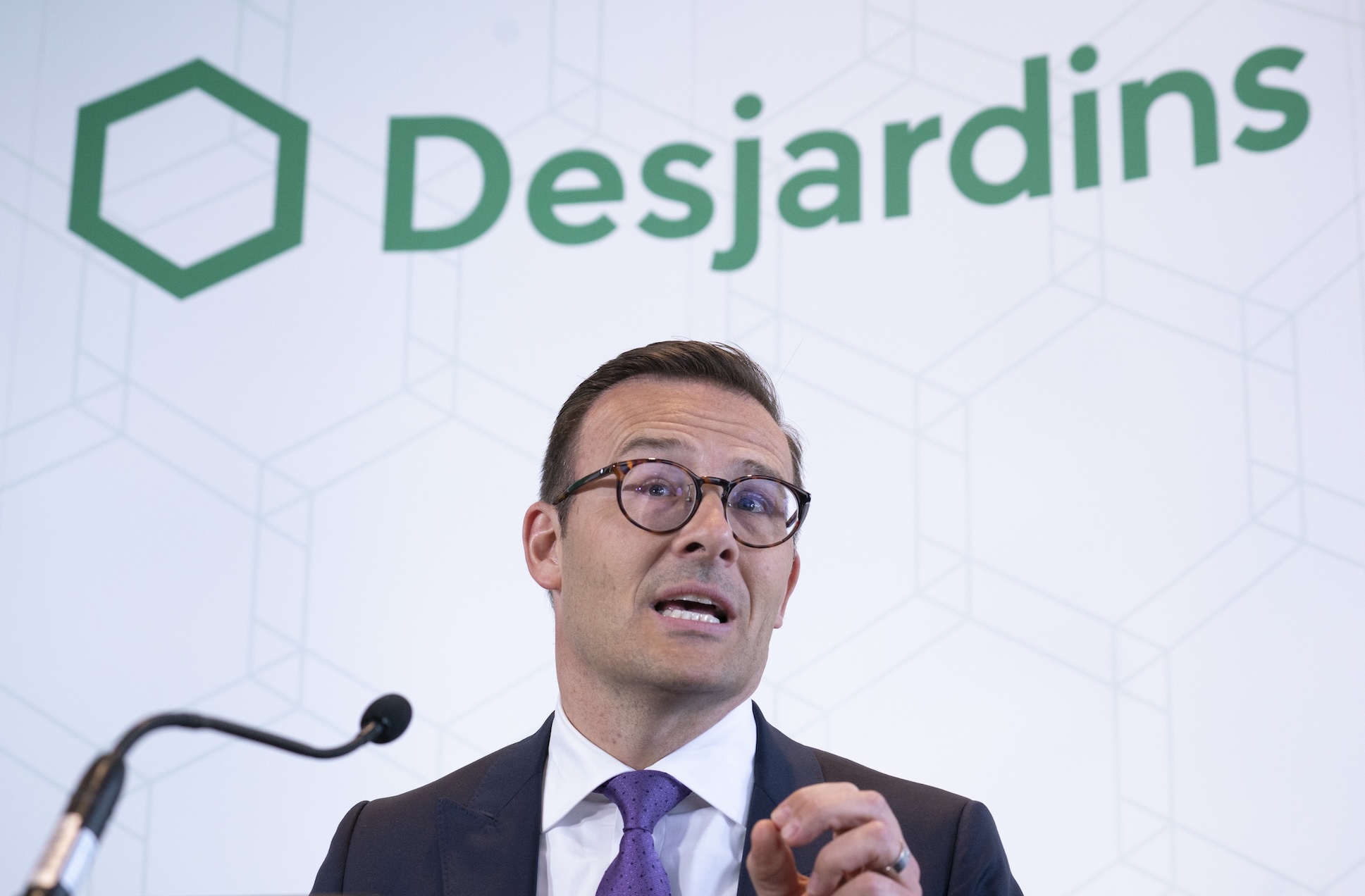 Desjardins president and CEO Guy Cormier gives a speech on stage at a news conference featuring a company banner in the background.