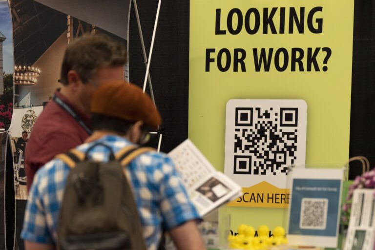 Two people talking at a job fair with a sign reading “Looking for work?” behind them.