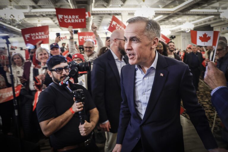 A shot of Mark Carney smiling as he greets Liberal supporters waving red signs with his name on them.
