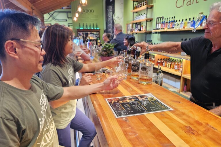 A shot of a bartender pouring whisky into a tasting glass held by guests standing along a bar made of laquered wood. There are three varieties of whisky in bottles on the bar in front of them.