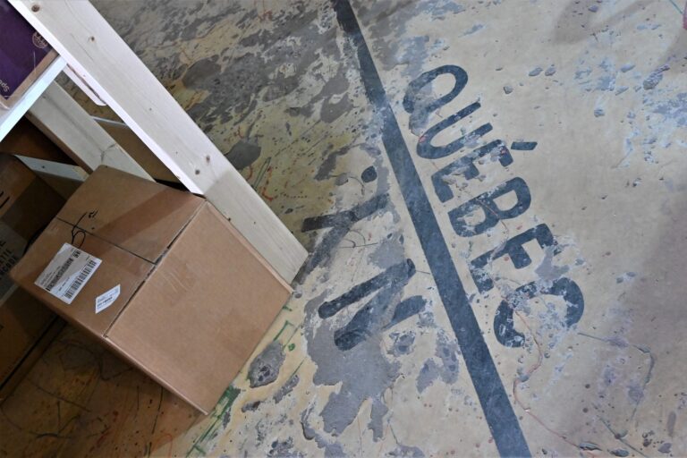 A parcel sits on a concrete floor with peeling paint beside a black line. The word "Québec" is painted on one side of the line; "N.Y." on the other.